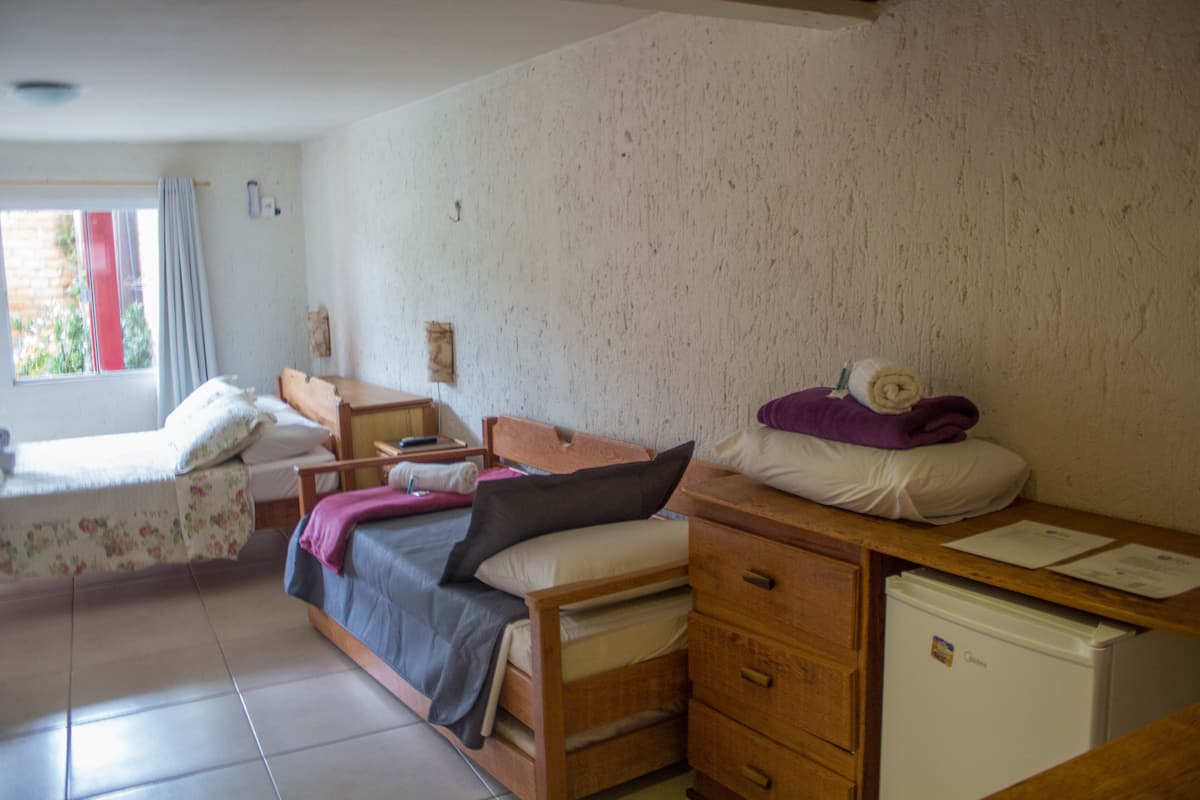Una habitación con dos camas, un aparador de madera, un refrigerador pequeño y una ventana con cortinas.