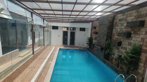 Una piscina interior con paredes de vidrio transparente, suelo de baldosas y algunas sillas de descanso.