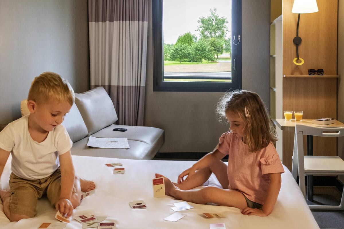 Zwei Kinder spielen mit Karten auf einem Bett in einem Hotelzimmer mit Blick auf Bäume durch das Fenster.