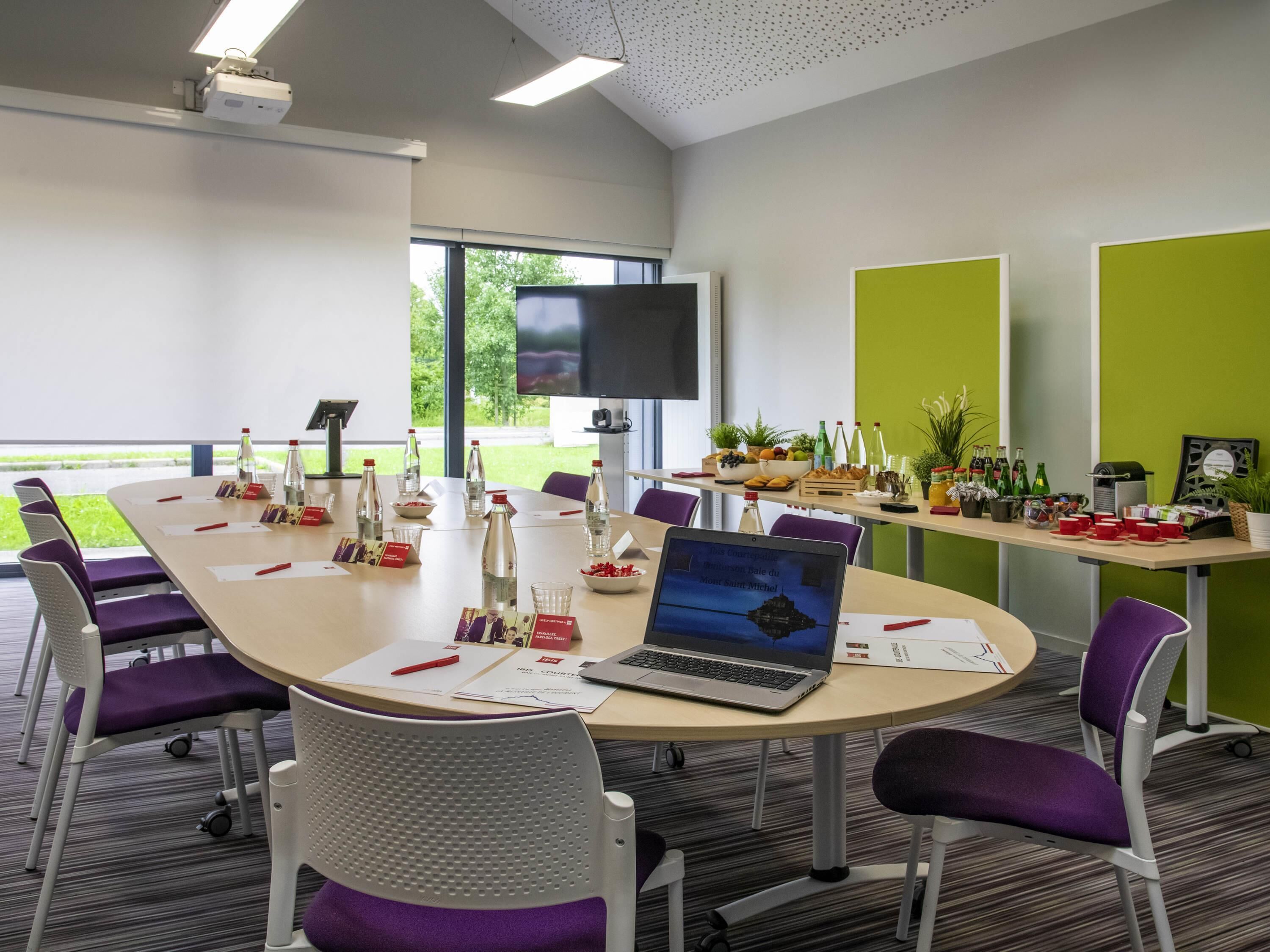 A conference room with a round table, chairs, a laptop, a TV, and a window with a view of greenery.