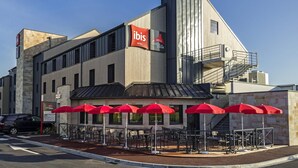 A modern building with a prominent 'ibis' sign, outdoor seating with red umbrellas, and a staircase leading to an upper level.