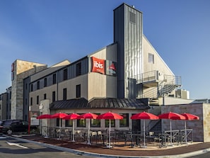 Un bâtiment moderne, reconnaissable à son grand panneau « ibis », doté d’une terrasse avec des parasols rouges et d’un escalier menant à un étage supérieur.