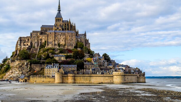 A historic castle with a prominent tower, situated on a rocky coastline with a beach in the foreground.
