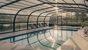 A covered swimming pool with a glass ceiling, lounge chairs, and a view of the surrounding landscape.