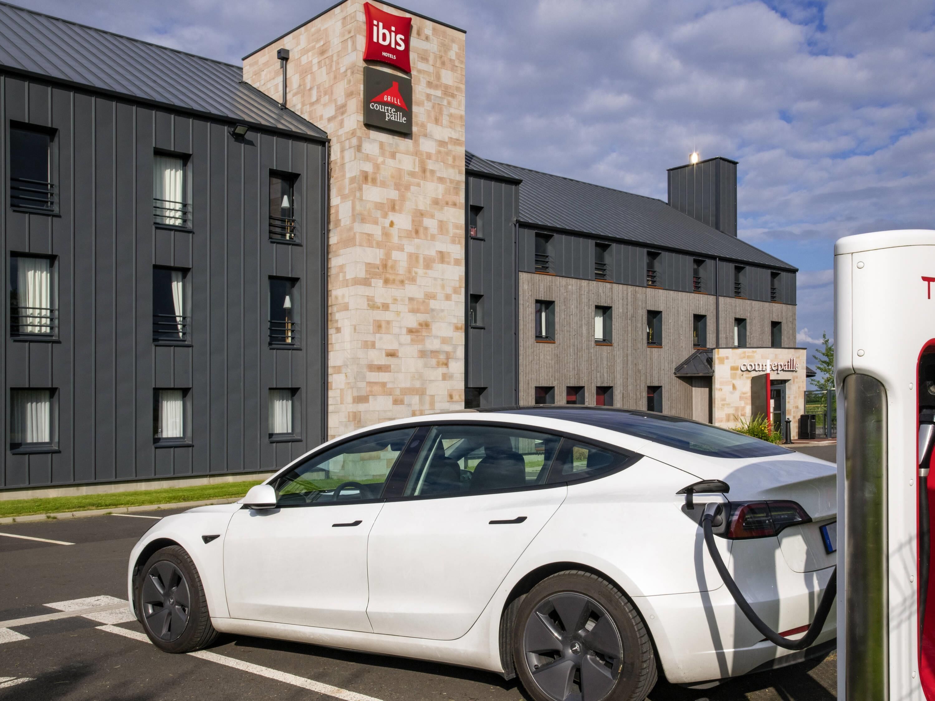 A white electric vehicle charging at a station in front of a hotel with a modern design, featuring a prominent sign for 'ibis Hotels' and 'Grill Courte Paillie'.