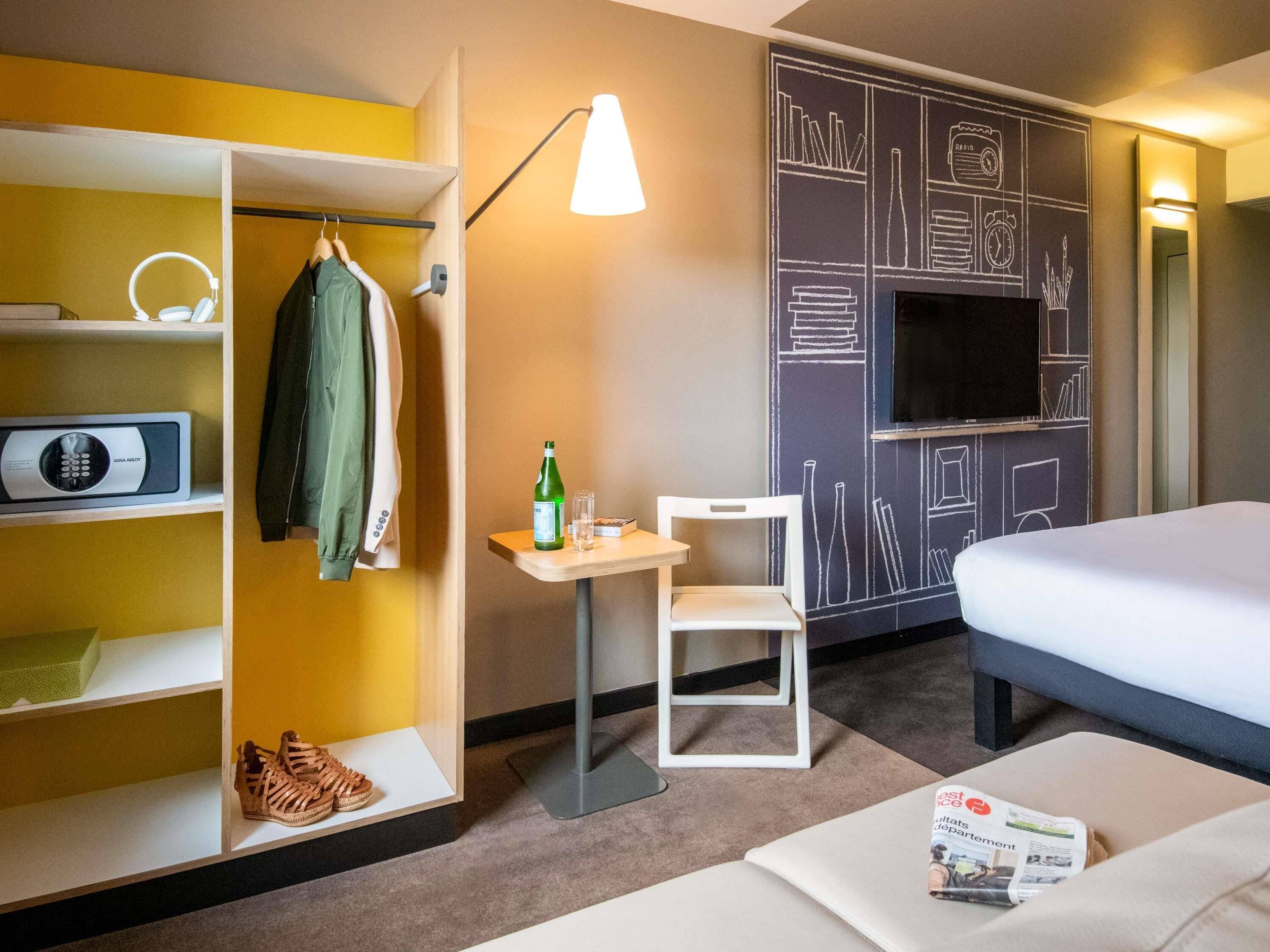A hotel room with a bed, a desk with a chair, a wardrobe with hanging clothes, a shoe shelf, a TV on the wall, and a lamp.