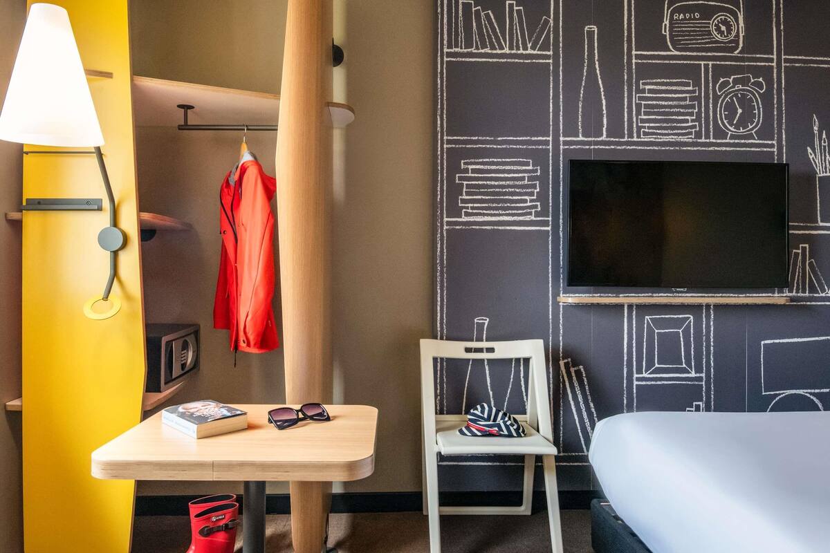 Ein Hotelzimmer mit Bett, Fernseher, Tisch mit Buch und Sonnenbrille, Stuhl mit Hut sowie Kleiderstange mit roter Jacke.