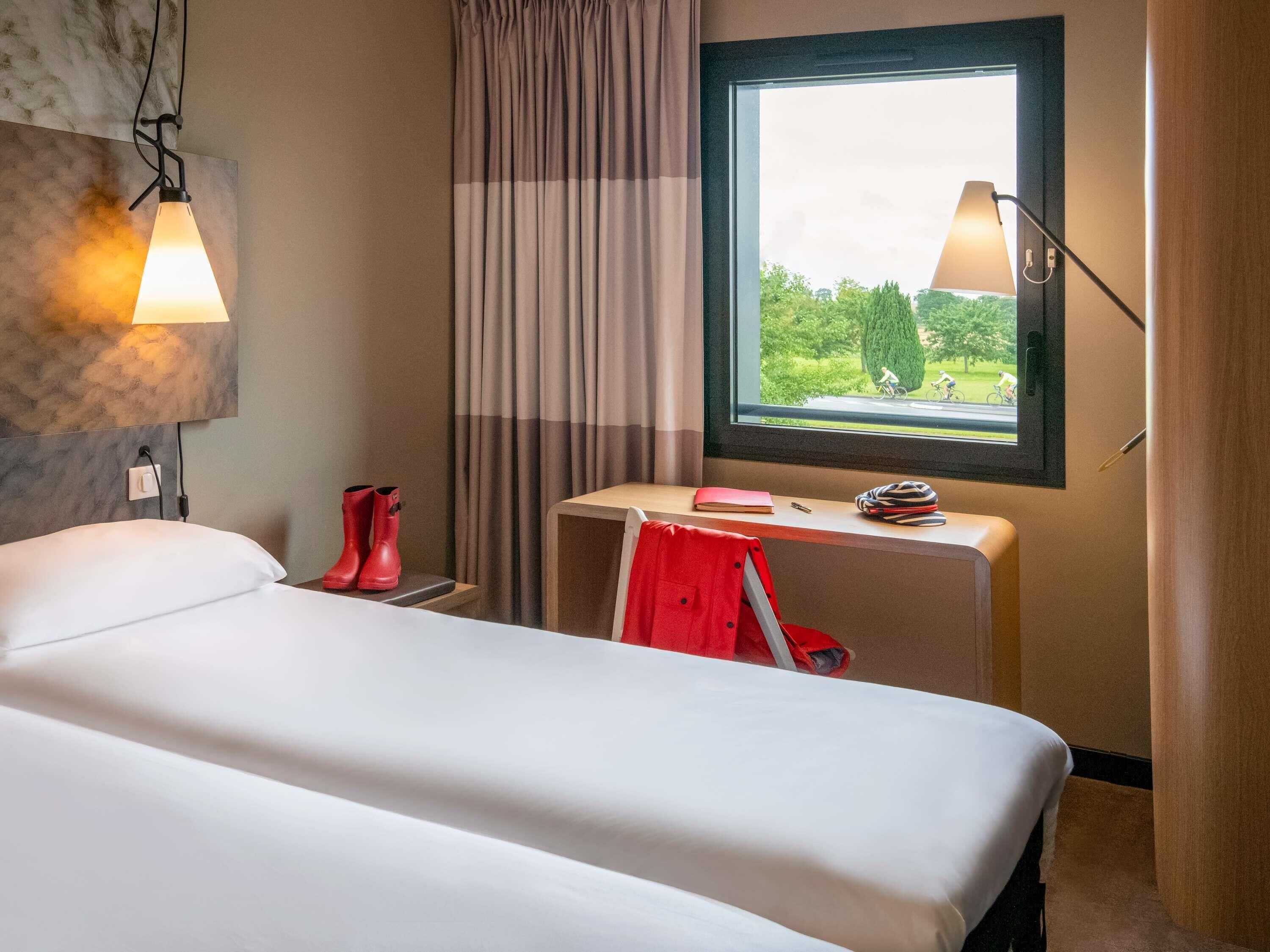 A hotel room with a bed, a desk with a chair, a window with a view of greenery, and a lamp.