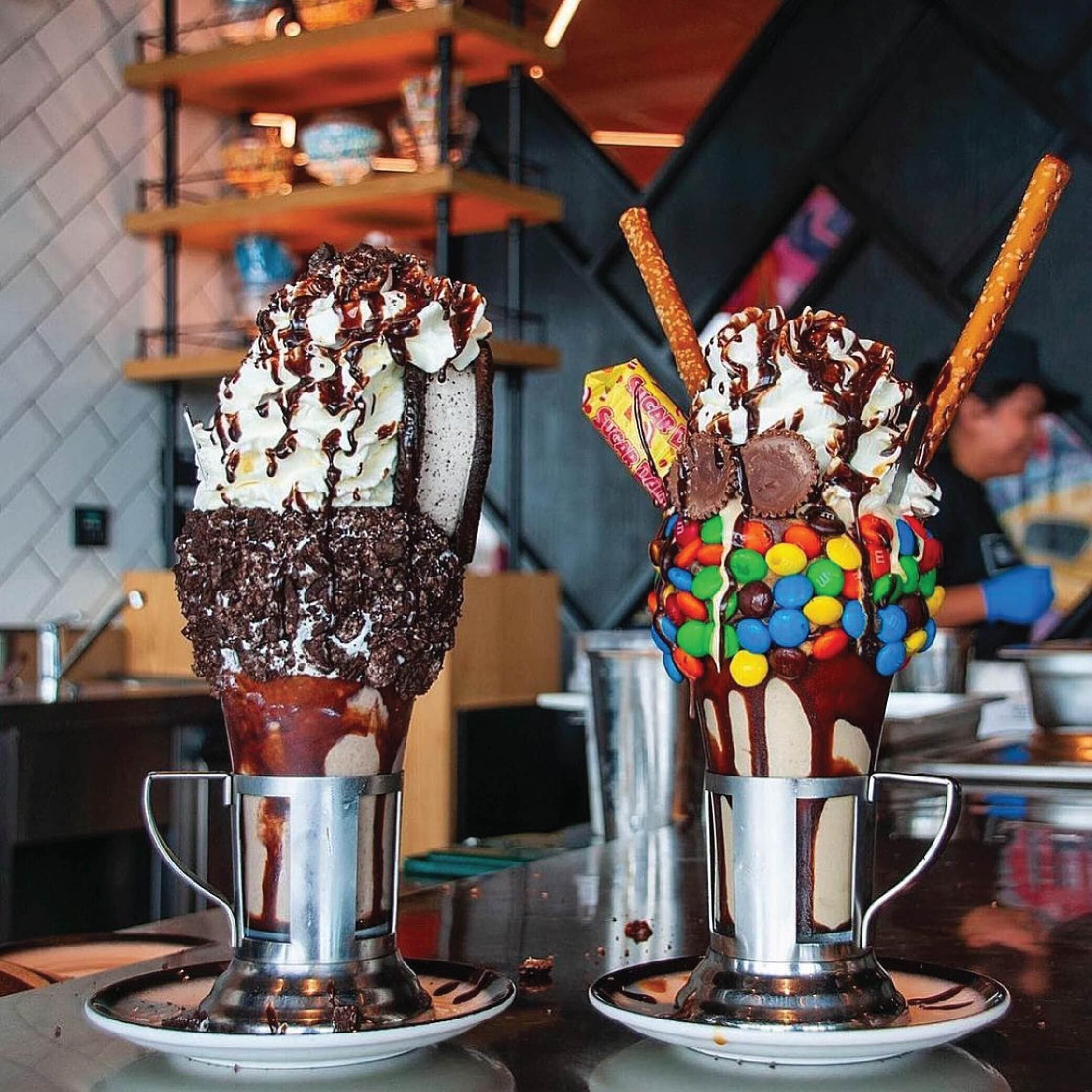 Two sundaes in metal cups with whipped cream, chocolate syrup, and toppings like candy and cookies.