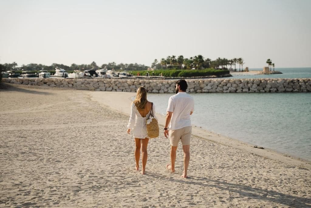 A couple walking on a sandy beach with a stone wall and palm trees in the background.