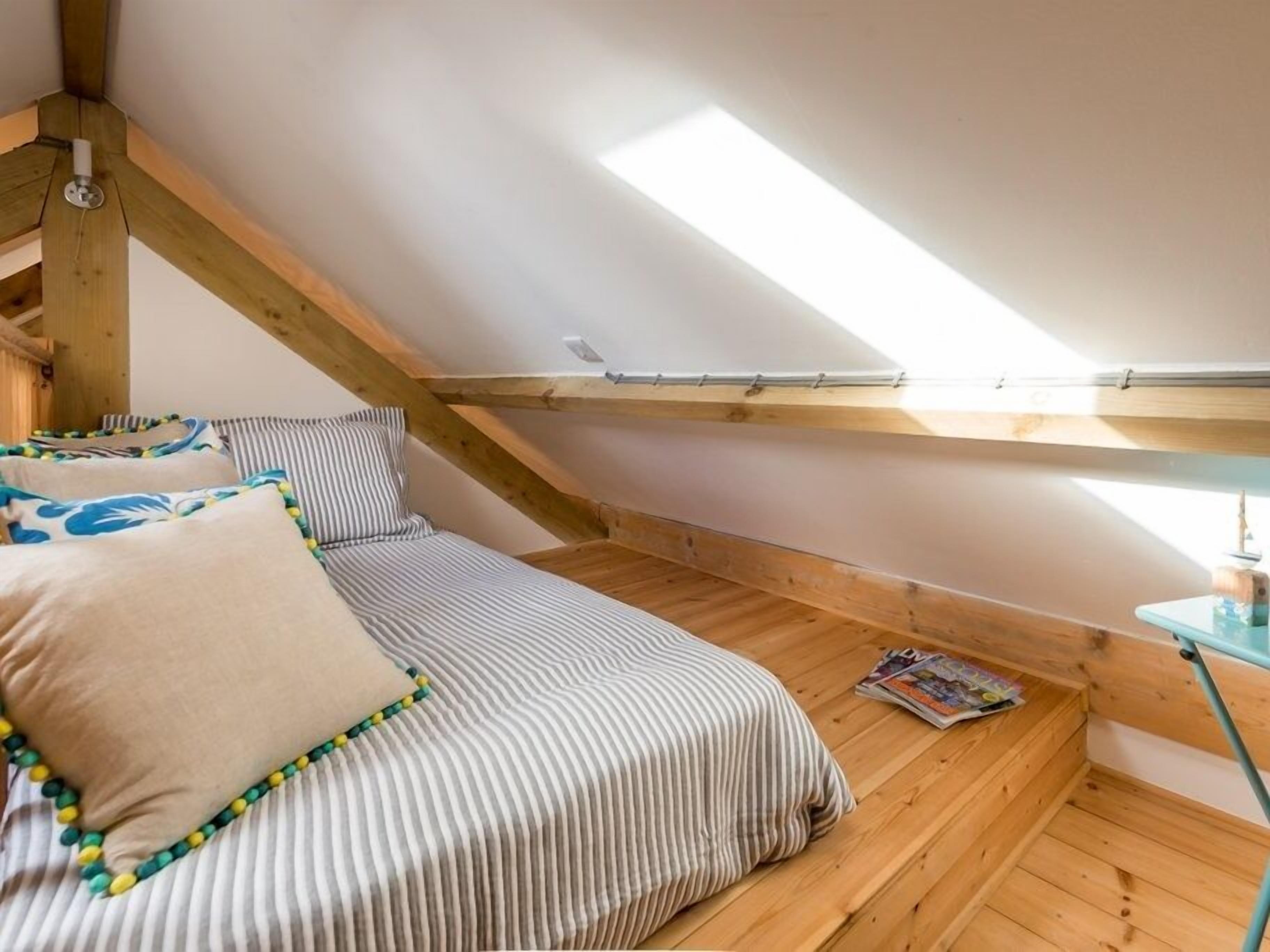Una piccola e accogliente camera da letto mansardata con tetto spiovente, travi in legno, un letto singolo con lenzuola a righe e un tavolino con una rivista.