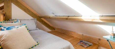 A small, cozy attic bedroom with a sloped ceiling, wooden beams, a single bed with striped bedding, and a small table with a magazine.