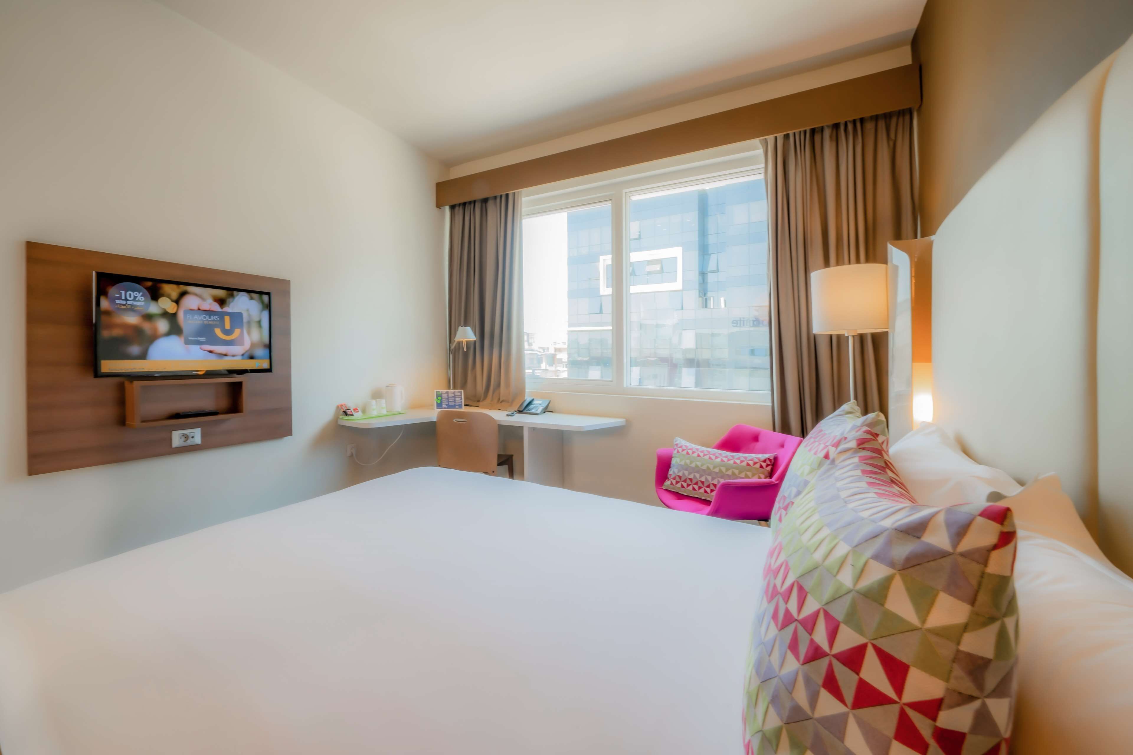 A hotel room with a large bed, a desk with a chair, a TV mounted on the wall, and a window with curtains.