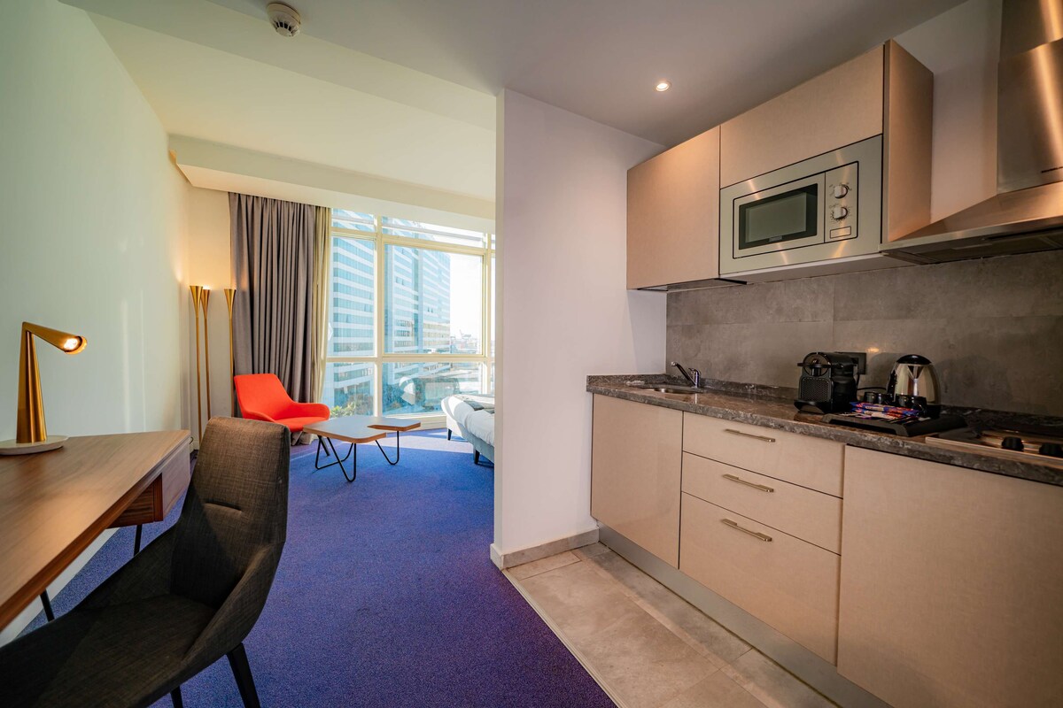 Habitación de hotel moderna con kitchenette, un comedor con mesa y sillas, y un ventanal con vistas a la ciudad.