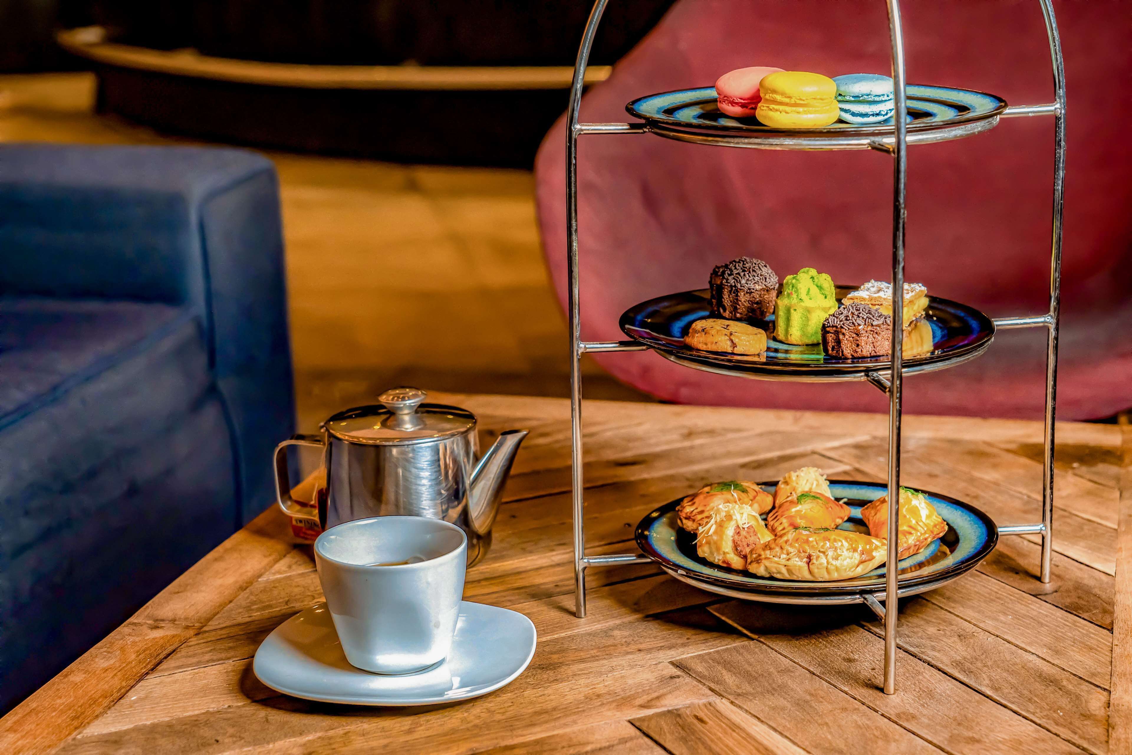 A three-tiered dessert stand with macarons and pastries on a wooden table, a teapot, and a cup of coffee.