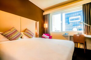 A hotel room with a large bed, colorful pillows, a desk with a chair, and a window with curtains.