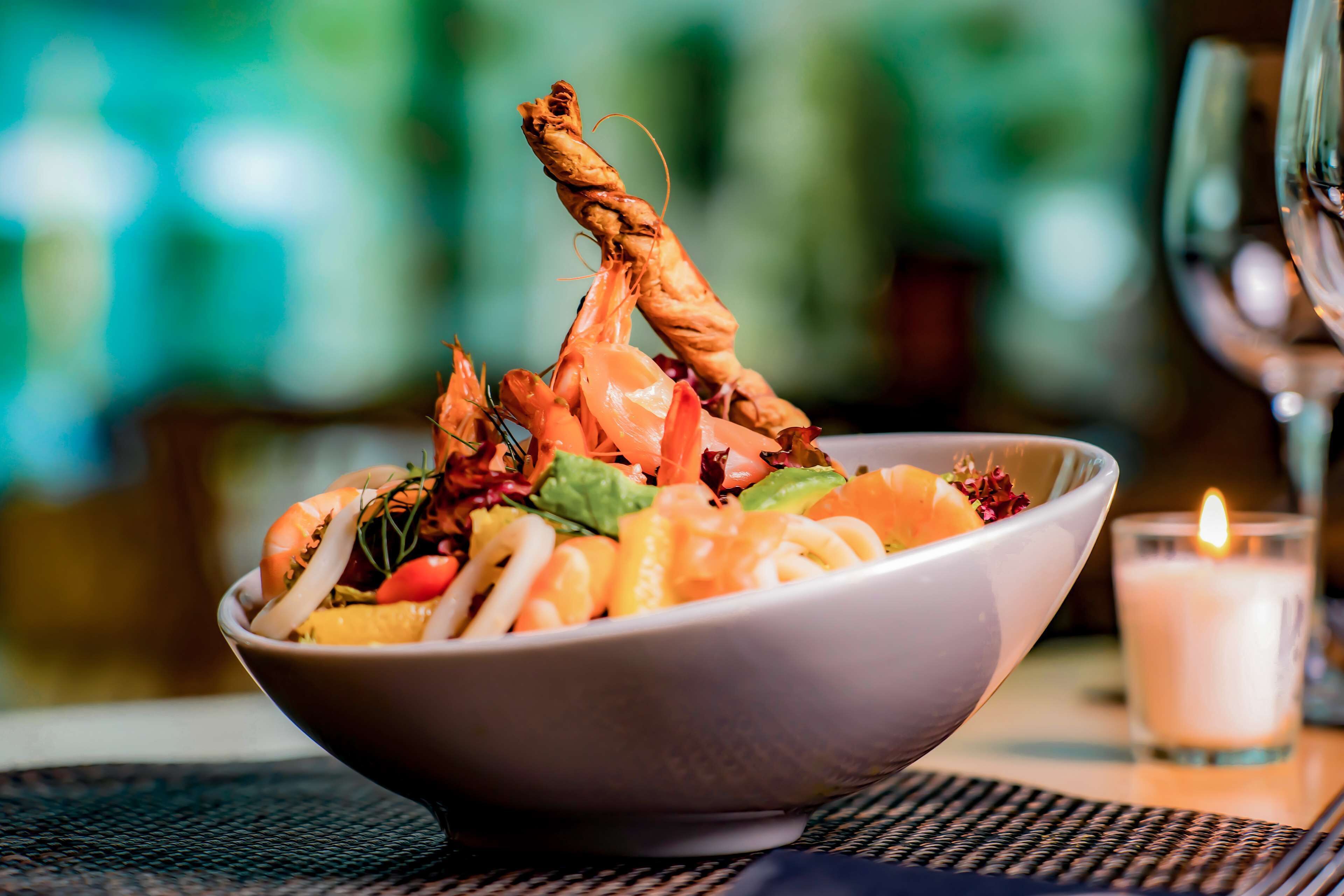 A bowl of seafood salad with shrimp and vegetables on a table with a candle.