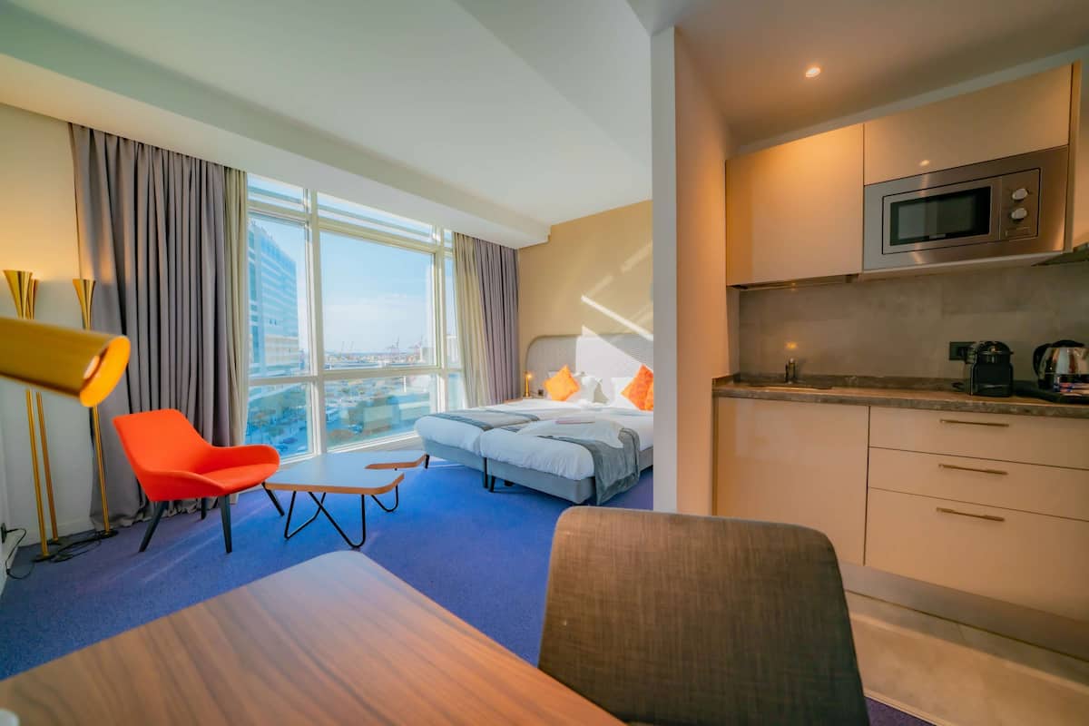 A hotel room with a bed, a desk, a chair, a kitchenette, and a large window with curtains.