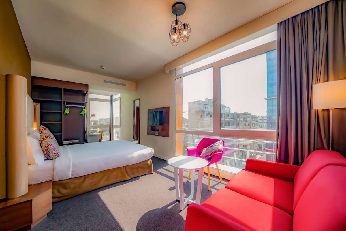 A modern hotel room with a bed, a red sofa, a small table, and a large window offering a city view.