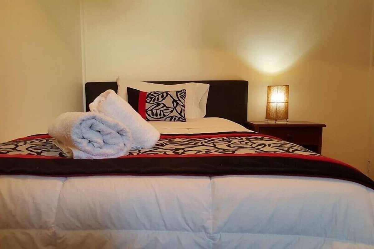 Una cama bien hecha con una toalla enrollada al lado, un cojín estampado y una lámpara de noche.