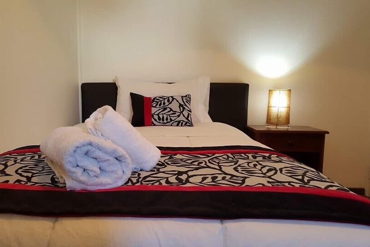Una cama bien hecha con una colcha estampada, un almohadón con diseño de hojas y una toalla enrollada sobre la cama.
