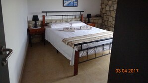 A bedroom with a bed, two bedside tables, a nightstand, a lamp, a painting, and a stone wall. -  exceptional holidays  (Gouvès Hersonissos Crête Grèce)