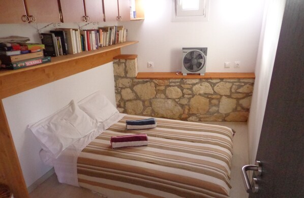 A bedroom with a bed, a stone wall, a fan, and shelves with books.
