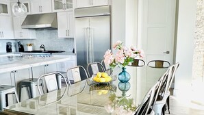 A modern kitchen with white cabinets, a stainless steel refrigerator, and a glass dining table with chairs.