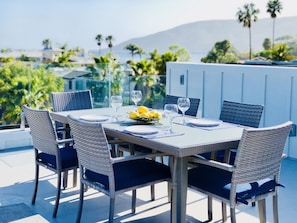 A dining table set with plates, glasses, and lemons, surrounded by wicker chairs with blue cushions, overlooking a scenic view with palm trees and a mountain in the background.