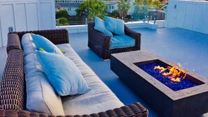 A rooftop terrace with wicker furniture, a fire pit, and a view of palm trees and buildings.