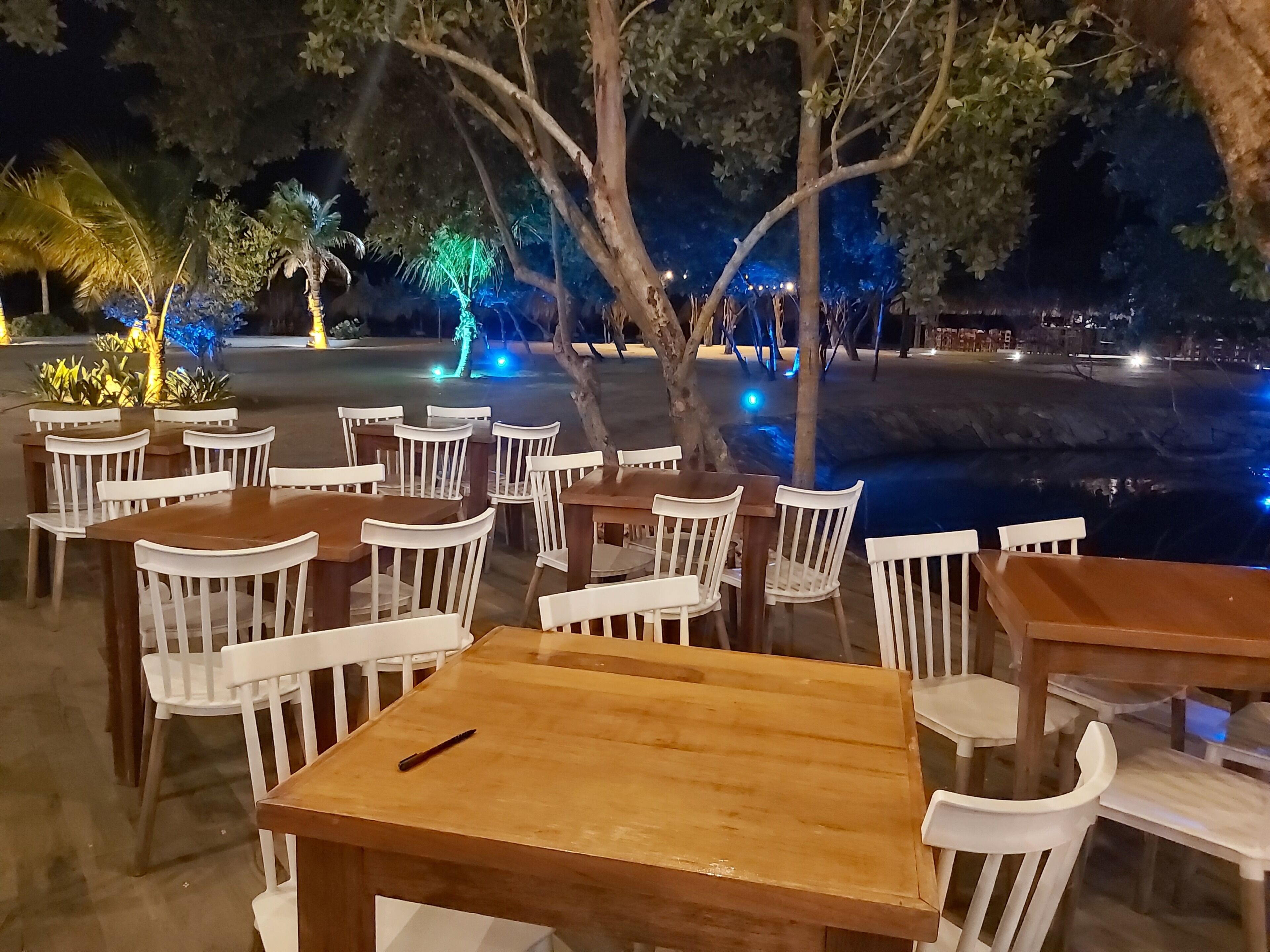An outdoor seating area with wooden tables and white chairs, surrounded by trees and illuminated by blue lights.