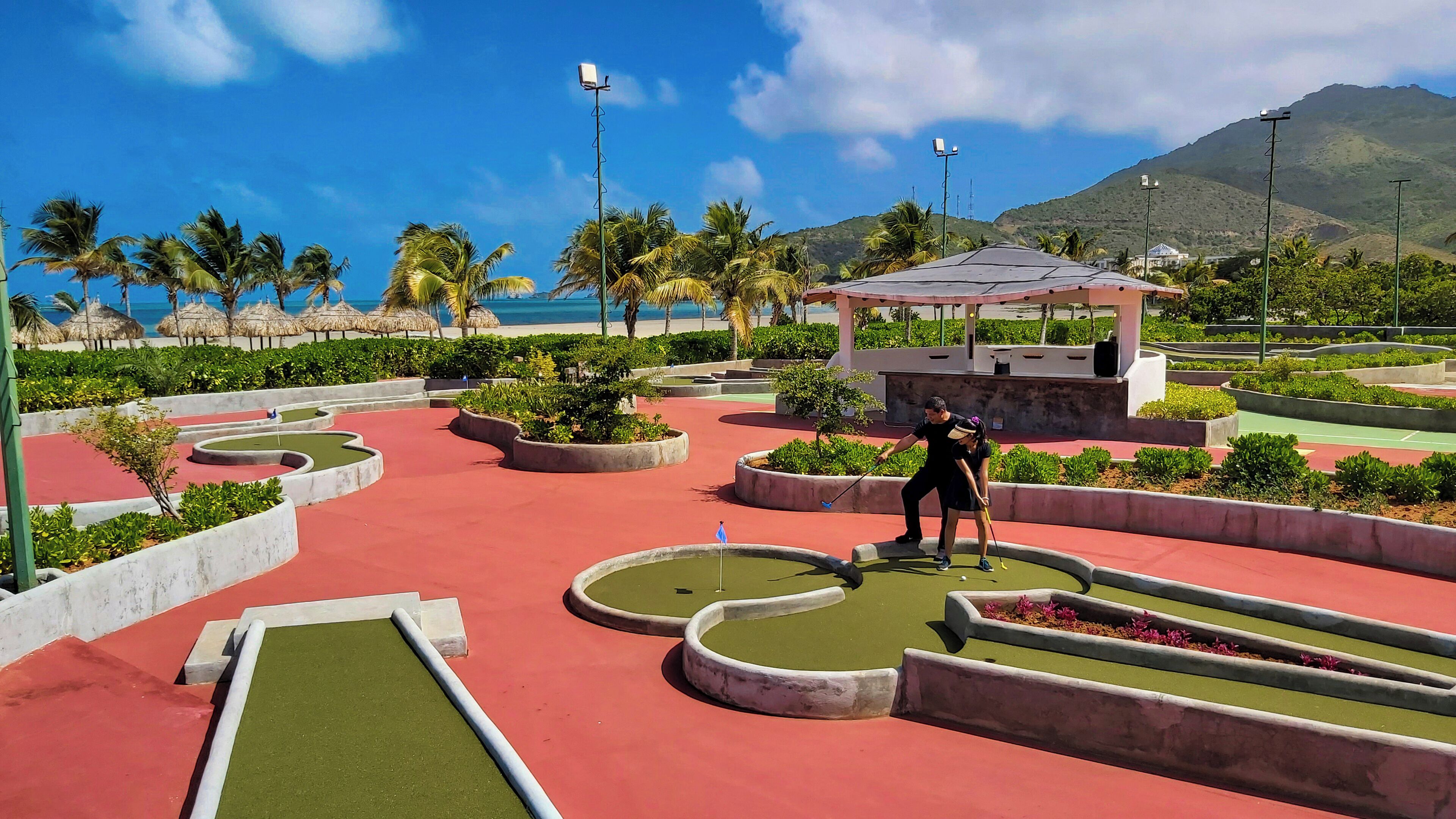A miniature golf course with a red and green playing surface, surrounded by landscaped areas and palm trees.