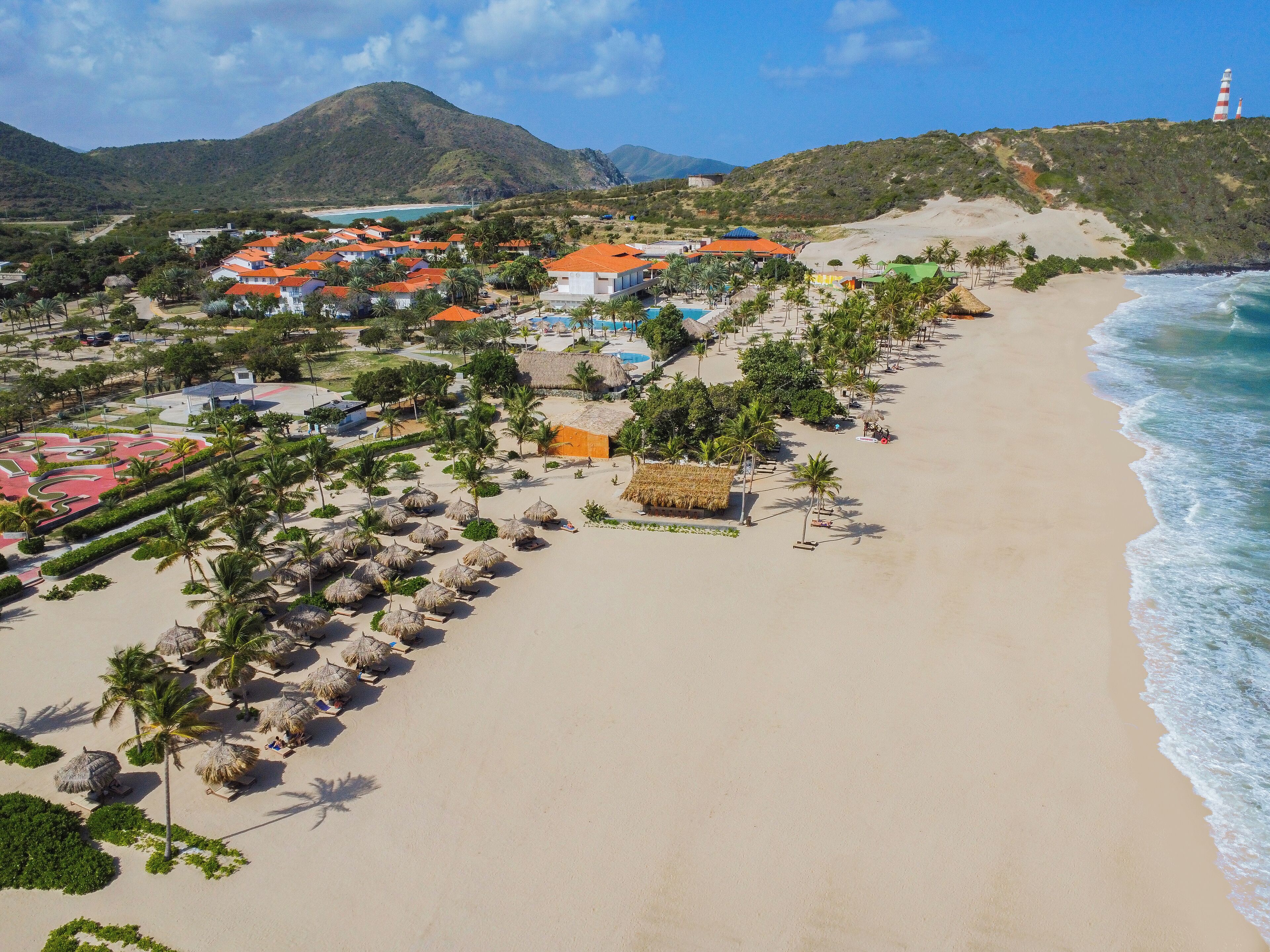 Un resort costero con cabañas frente a la playa, palmeras y montañas al fondo.