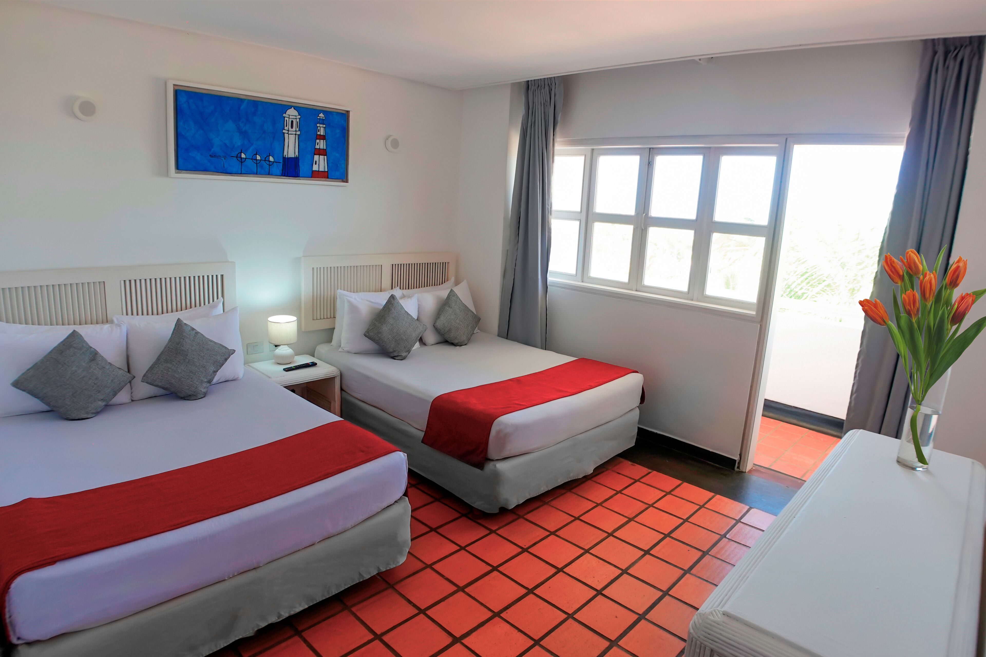 Camera d'albergo con due letti, un comodino, una lampada, un quadro, una finestra con tende e un vaso con fiori.
