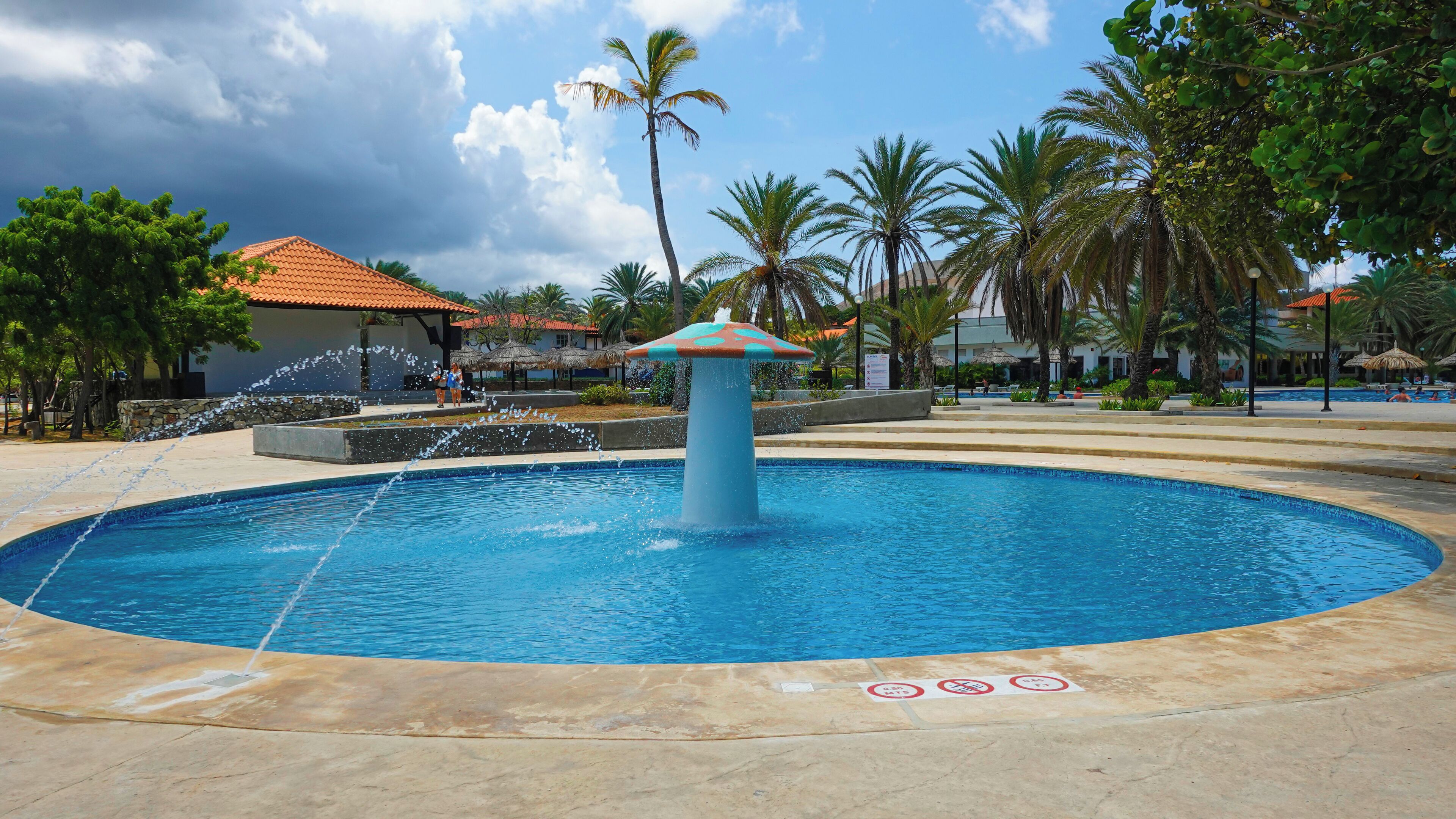 A swimming pool with a central fountain, surrounded by palm trees and a building with a red-tiled roof.