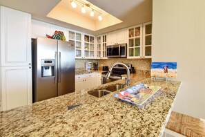 A modern kitchen with stainless steel appliances, a granite countertop, and a painting of a beach scene. - Coastal chic, updated tile, beachfront condo w/balcony! 🐚 (Destin)