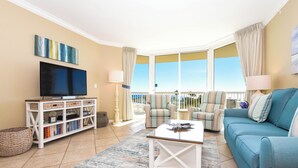 Sala de estar com televisão, sofá, poltronas e varanda com cortinas.