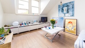 A modern, well-lit room with a white desk, a white dining table, and a large window. There are various decorative items on the desk, including a vase with flowers, a bowl, and a small plant. The walls are adorned with artwork, and there is a television on a white cabinet. - Lofty Ideals (London)