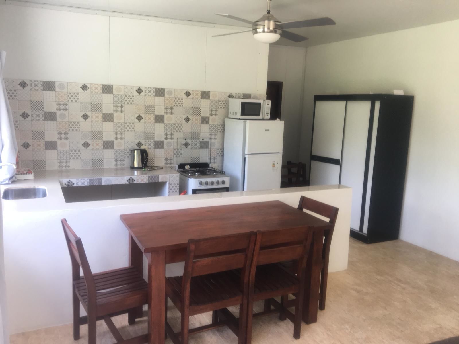 A compact kitchen with a gas stove, microwave, and refrigerator. There is a dining table with chairs and a ceiling fan above.