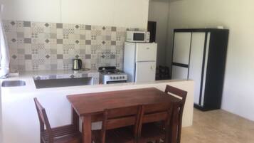 A compact kitchen with a gas stove, microwave, and refrigerator. There is a dining table with chairs and a ceiling fan above.