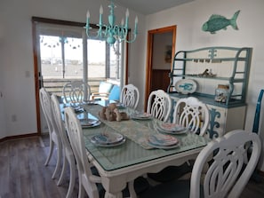 A dining area with a glass table, white chairs, and a chandelier. There is a fish decoration on the wall and a wooden door.