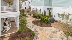A residential area with a paved walkway, small trees, and a wooden deck with chairs.