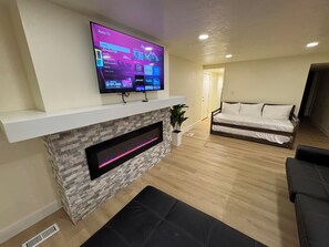 A modern living room with a flat-screen TV mounted above a fireplace, a sofa, and a bed in the background. - Fast WiFi | Foosball | Close to Conference Centers. (Midvale)