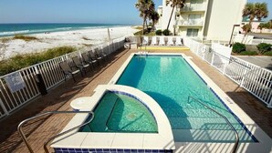 Una piscina rectangular con un trampolín, rodeada por una terraza con sillas y una cerca blanca, con vistas a una playa y al océano.