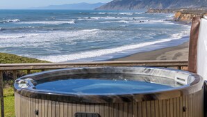 Un jacuzzi en una terraza de madera con vistas a una playa, olas y una ladera boscosa al fondo.