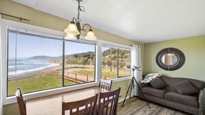 Una habitación con una mesa de comedor y sillas, un sofá, un trípode y un amplio ventanal con vistas a la playa y las colinas.