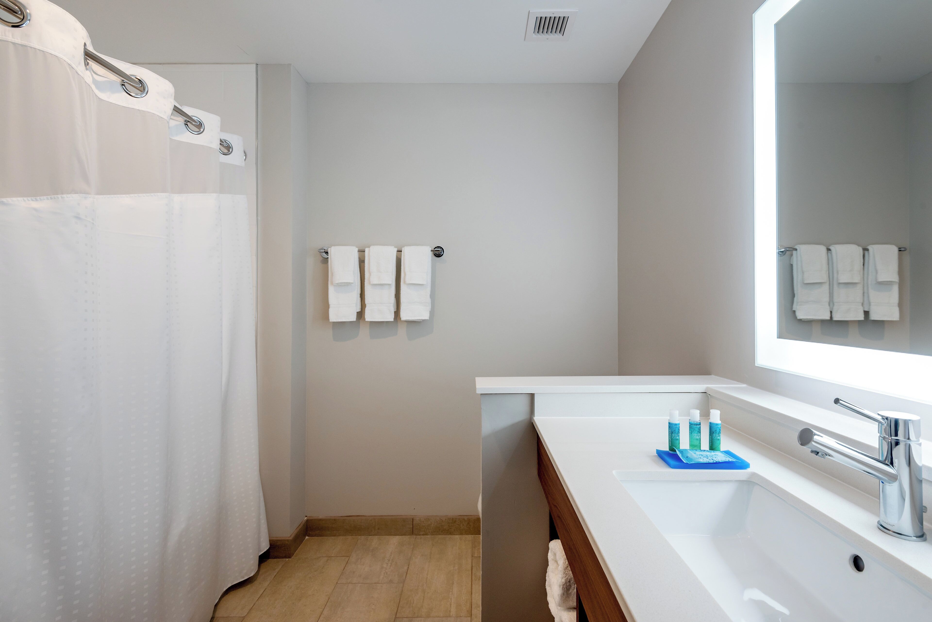 A bathroom with a shower curtain, three towels on a rack, a sink with a mirror, and toiletries on the counter.