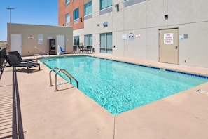 A clear swimming pool with a lifeguard chair and a safety sign, a covered seating area, and a building with a fire alarm control panel. - Holiday Inn Express & Suites El Paso East-Loop 375 by IHG (El Paso)