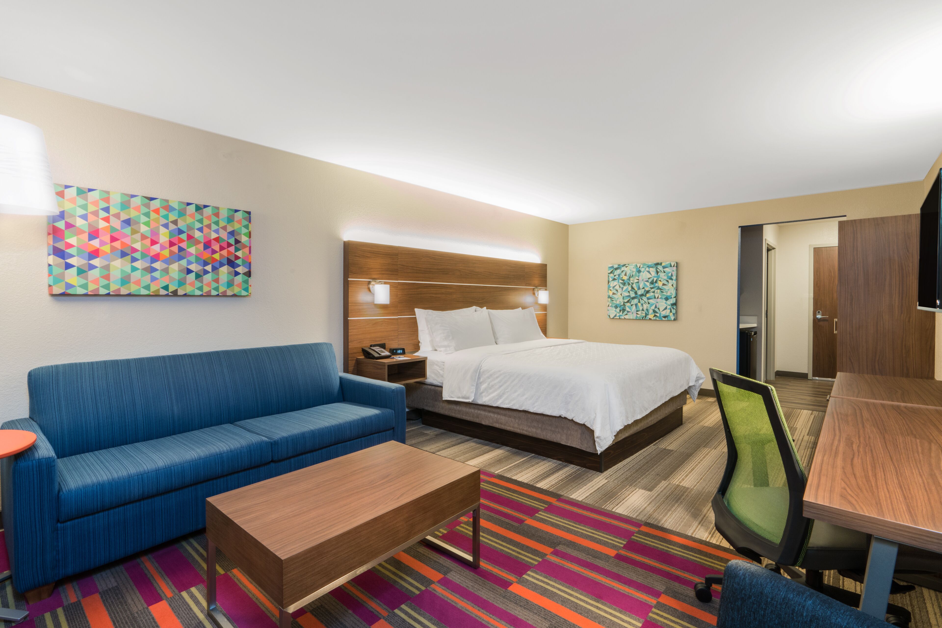 A hotel room with a bed, a blue sofa, a wooden coffee table, a colorful striped rug, and a desk with a chair.