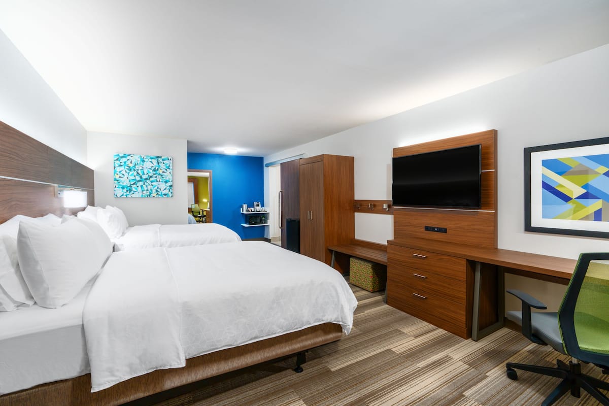 Une chambre d’hôtel équipée d’un lit, d’un bureau, d’une chaise, d’une télévision et décorée avec des œuvres d’art aux murs.
